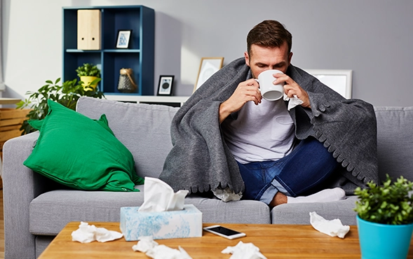 Erkälteter Mann auf dem Sofa mit Tasse und umgeben von benutzten Taschentüchern